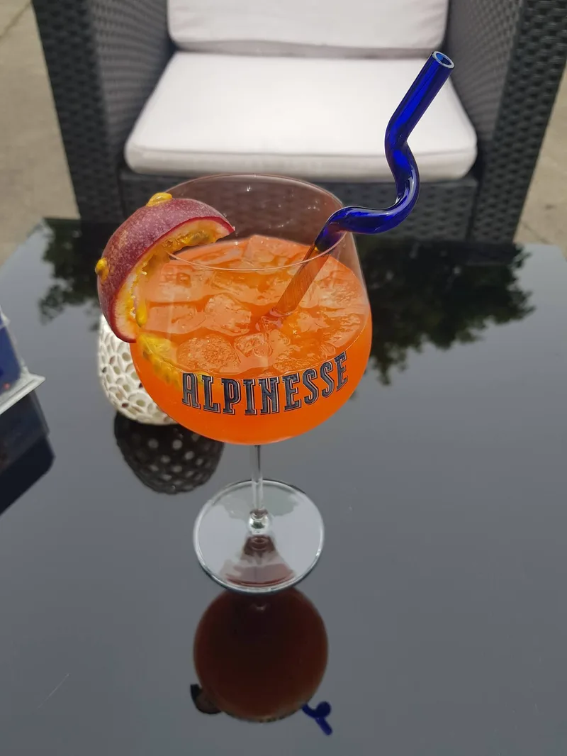 Ein Aperol im bauchigen Alpinesse-Glas mit Eis, einer halben Passionsfrucht am Rand und einem blauen Glastrinkhalm, auf einem Tisch der Terrasse.