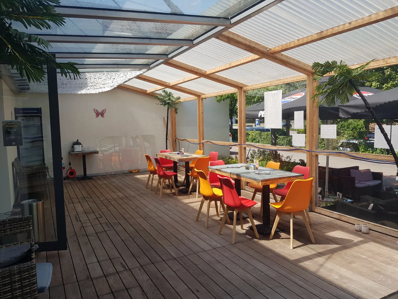 Die überdachte Terrasse mit Holzboden, zwei Tischen und roten und orangen Stühlen; das Dach ist teils Glas, teils Wellplatte.