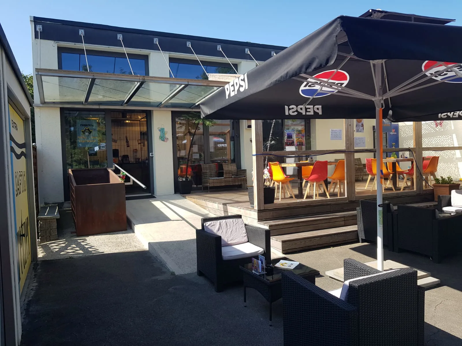 Der Vorplatz des Pulfer-fass: links der Eingang mit Glasvordach, rechts eine überdachte Holzterrasse mit roten und orangen Stühlen, davor Loungesessel unter einem Sonnenschirm.
